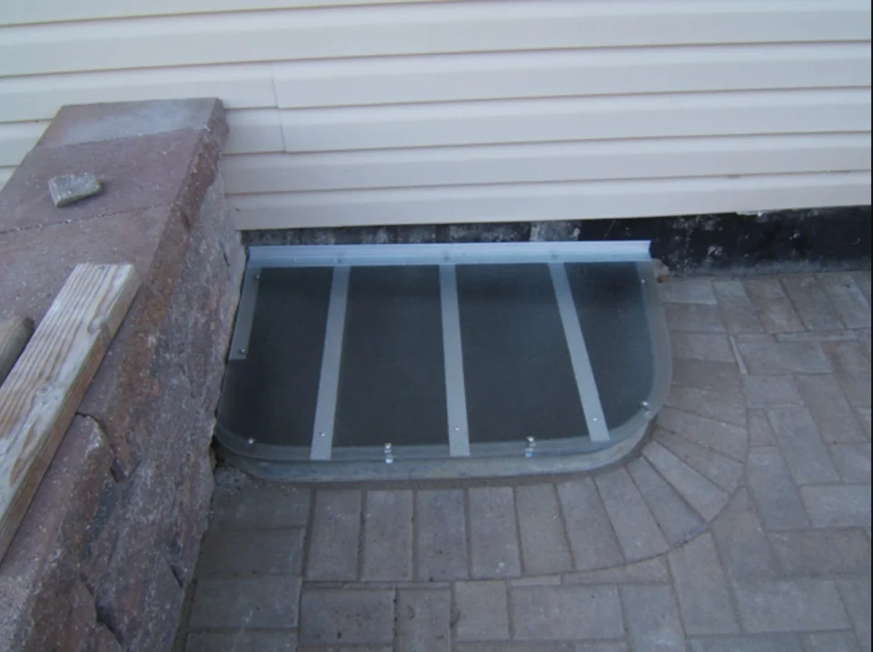 Rectangular custom window well cover on a paver patio next to cream siding