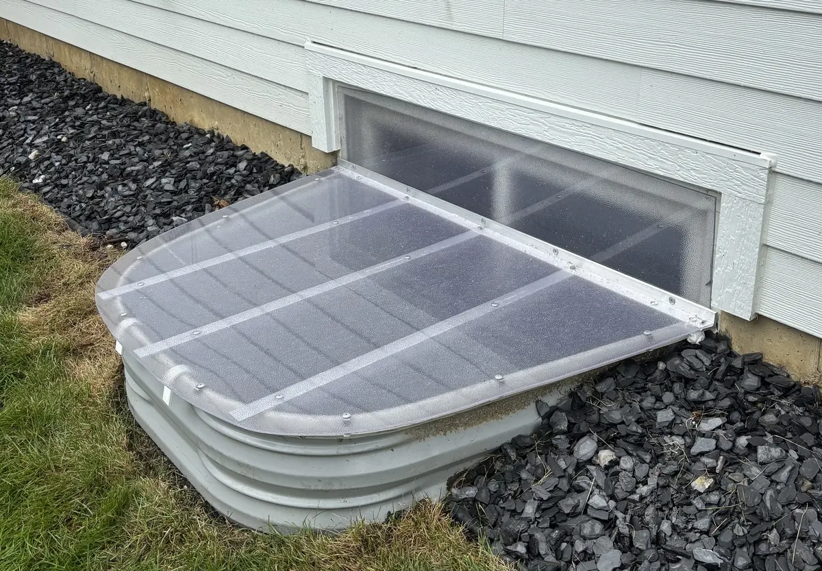 Clear window well cover installed over a galvanized well on a home with siding and rock landscaping